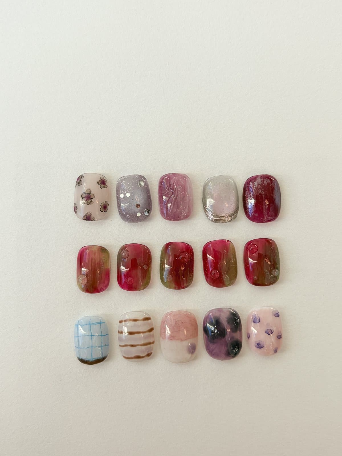 Fifteen October nail art sample tips in three rows in even light, in burgundy, deep pink, milky pink, plum, fine brown stripes and a pale blue check, by Joy Nail Studio Vancouver